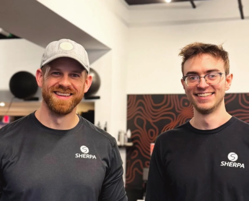 Two Sherpa fitness trainers smiling and wearing branded shirts at the gym, showcasing the friendly, personalized coaching experience that helps clients build strength and confidence beyond the gym.