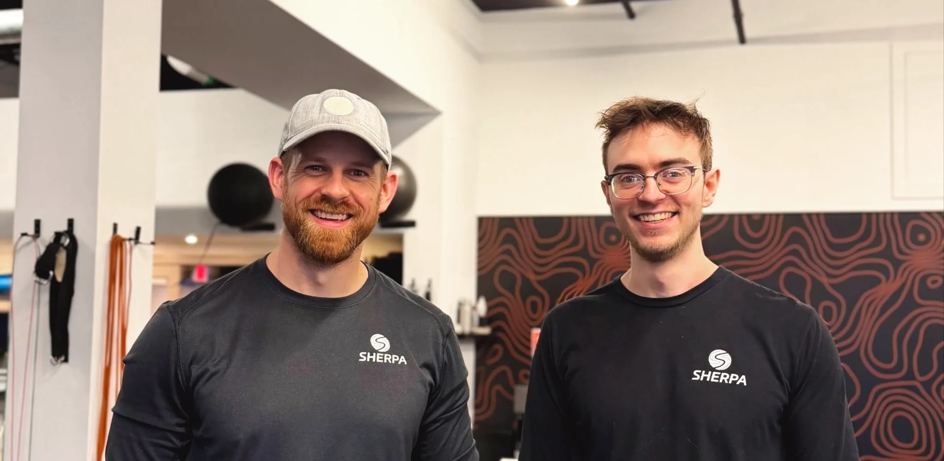Two Sherpa fitness trainers smiling and wearing branded shirts at the gym, showcasing the friendly, personalized coaching experience that helps clients build strength and confidence beyond the gym.